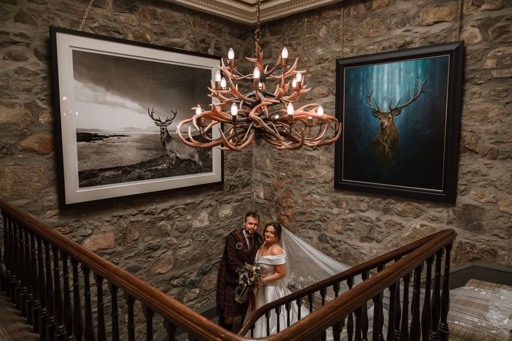 Bride and groom under antler chandelier on stone staircase with Highland stag artwork in Scottish wedding venue.