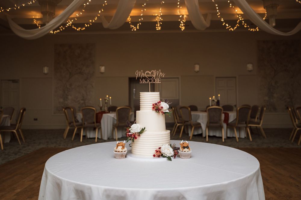 Elegant four-tier white wedding cake on draped table in softly lit reception venue with string lights.