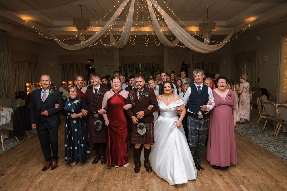 Bride and groom lead wedding guests in joyful dance on decorated ballroom floor at Scottish wedding reception.