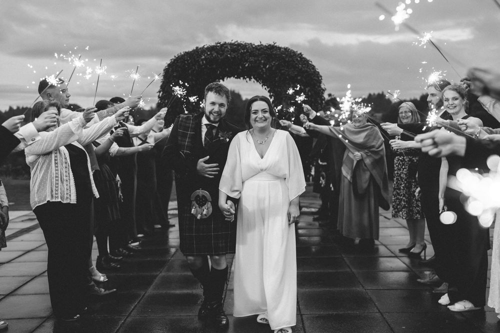 Bride and groom walk through sparkler tunnel at wedding, guests celebrating outdoors in black and white photo.
