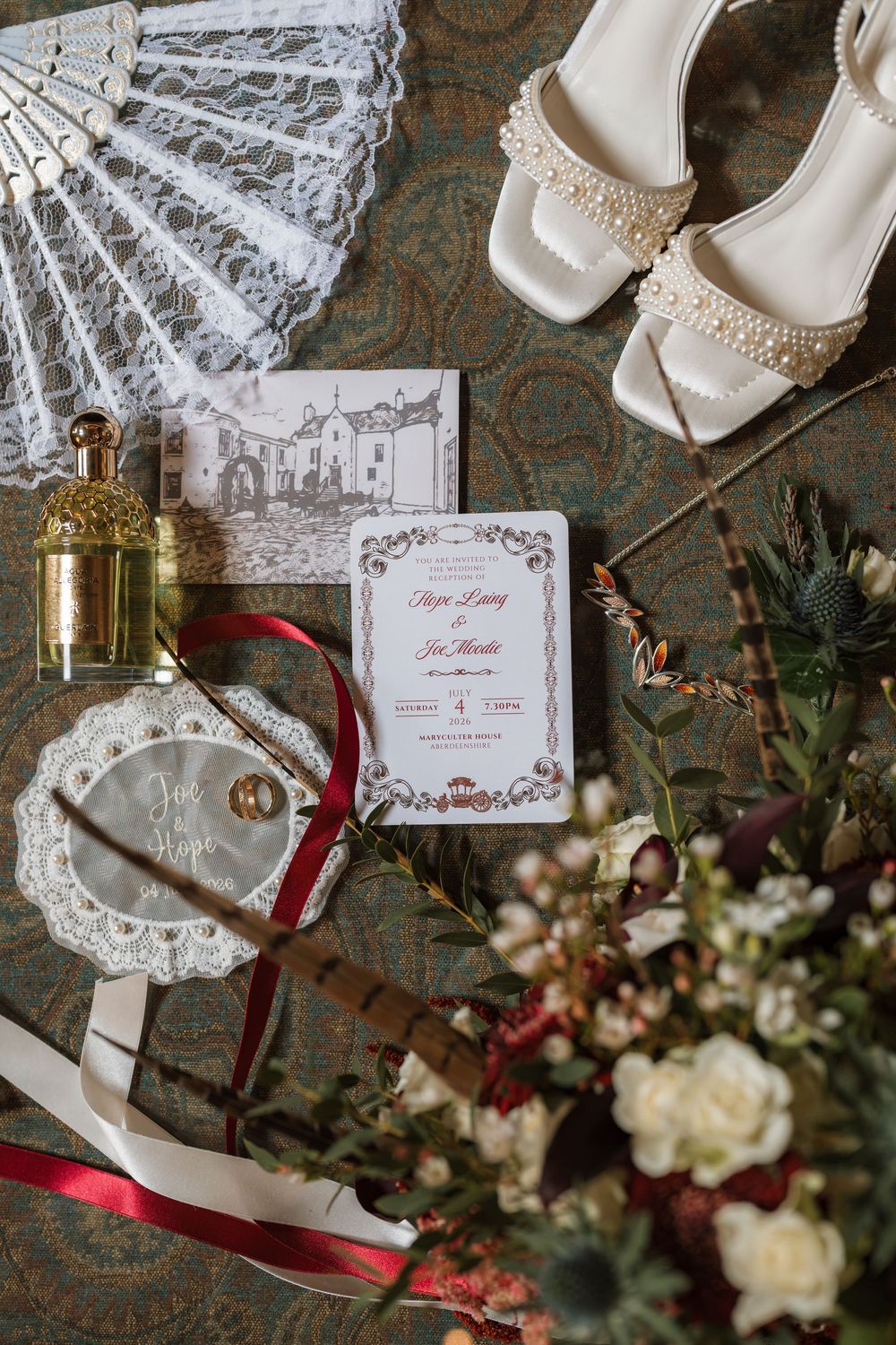 Elegant wedding flat lay with pearl heels, lace fan, floral invitation, gold perfume, rings, red ribbons, and bridal bouquet.