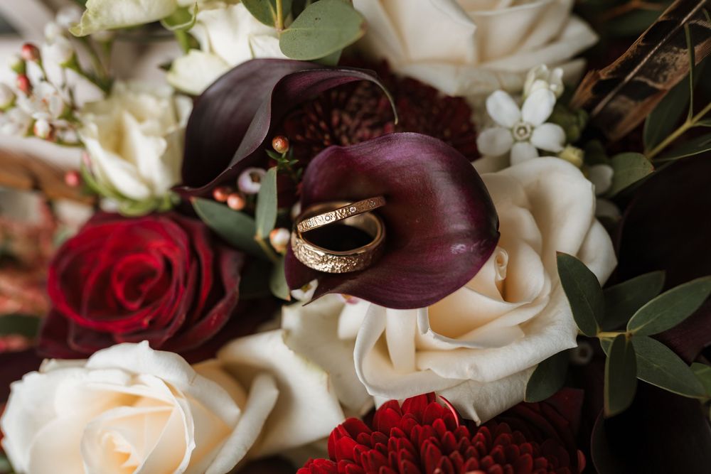 Gold wedding rings resting on a lush bridal bouquet of burgundy calla lilies, red and ivory roses.