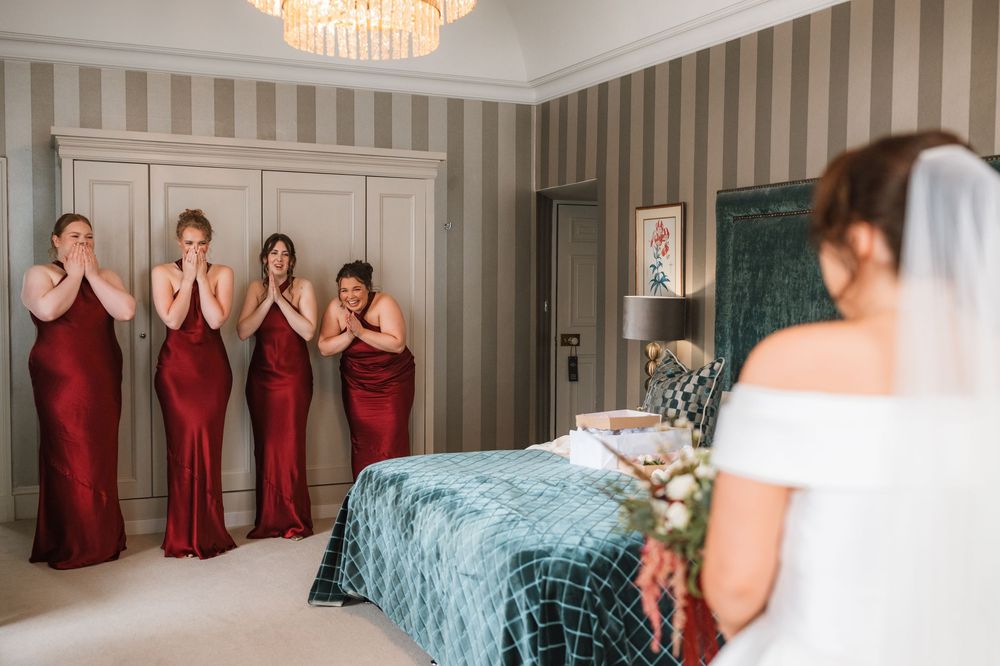 Bridesmaids in red gowns react emotionally as bride in white wedding dress and veil reveals her look in elegant bedroom.