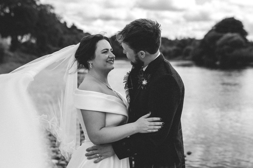 Black and white photo of a smiling bride and groom embracing by a lake on their wedding day.