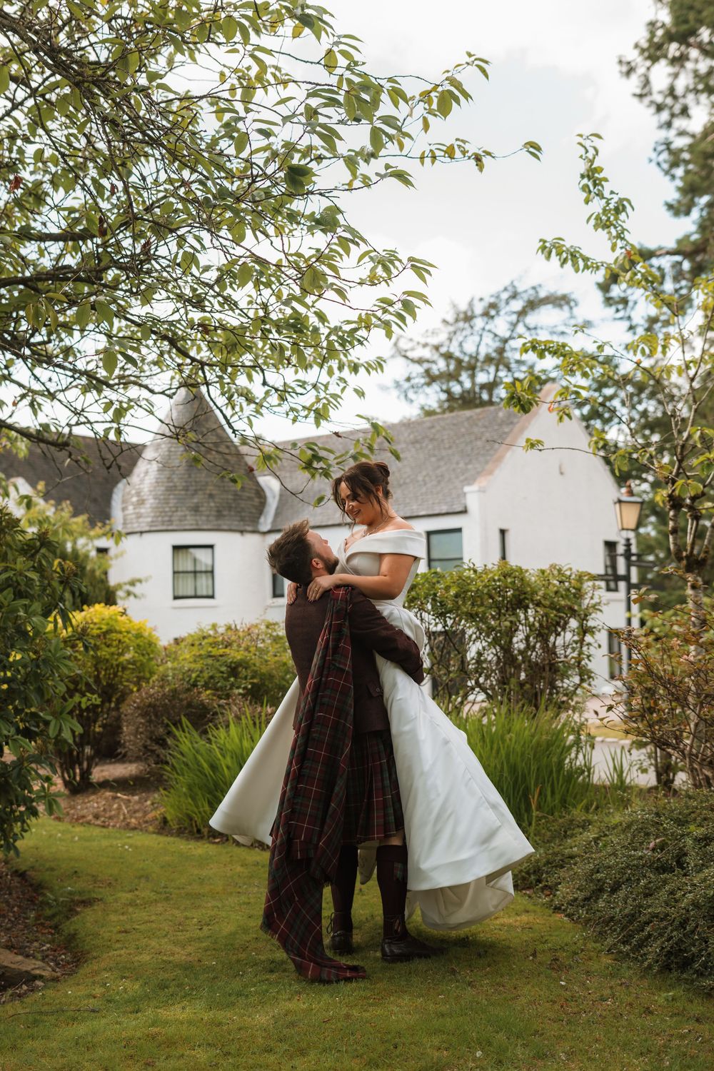 Groom in tartan kilt lifting bride in white gown in lush garden outside white Scottish country house.