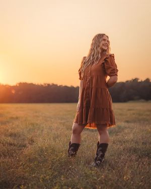 Joyful young woman in brown dress and cowboy boots laughs in open field during vibrant sunset.