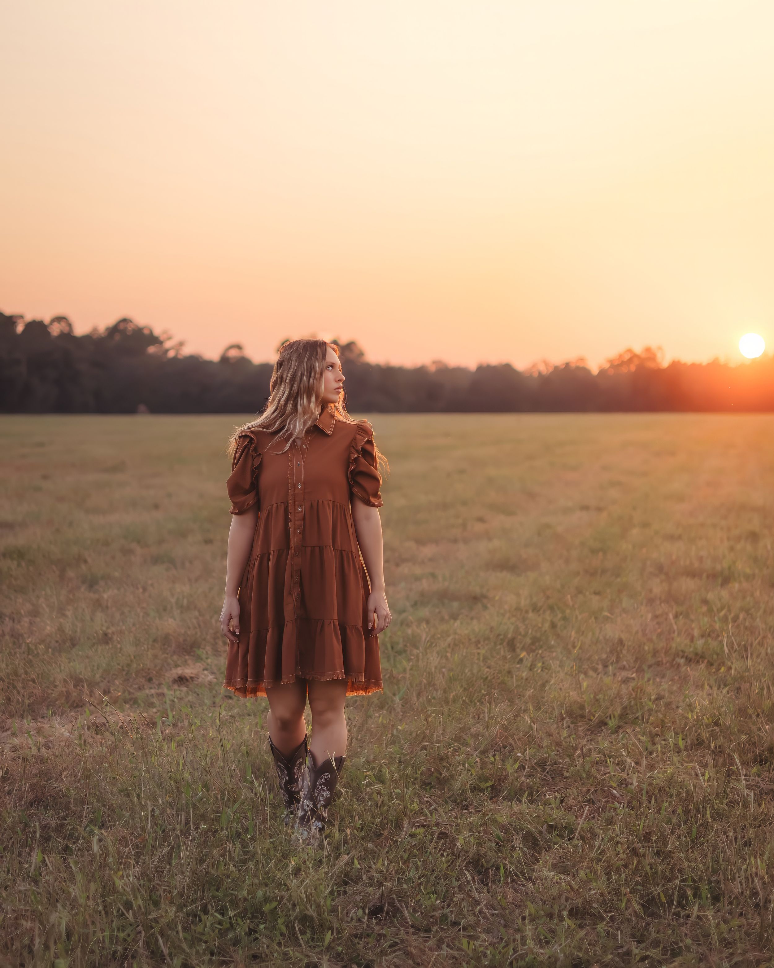 Young woman in rust brown dress and cowboy boots stands in golden field at sunset.