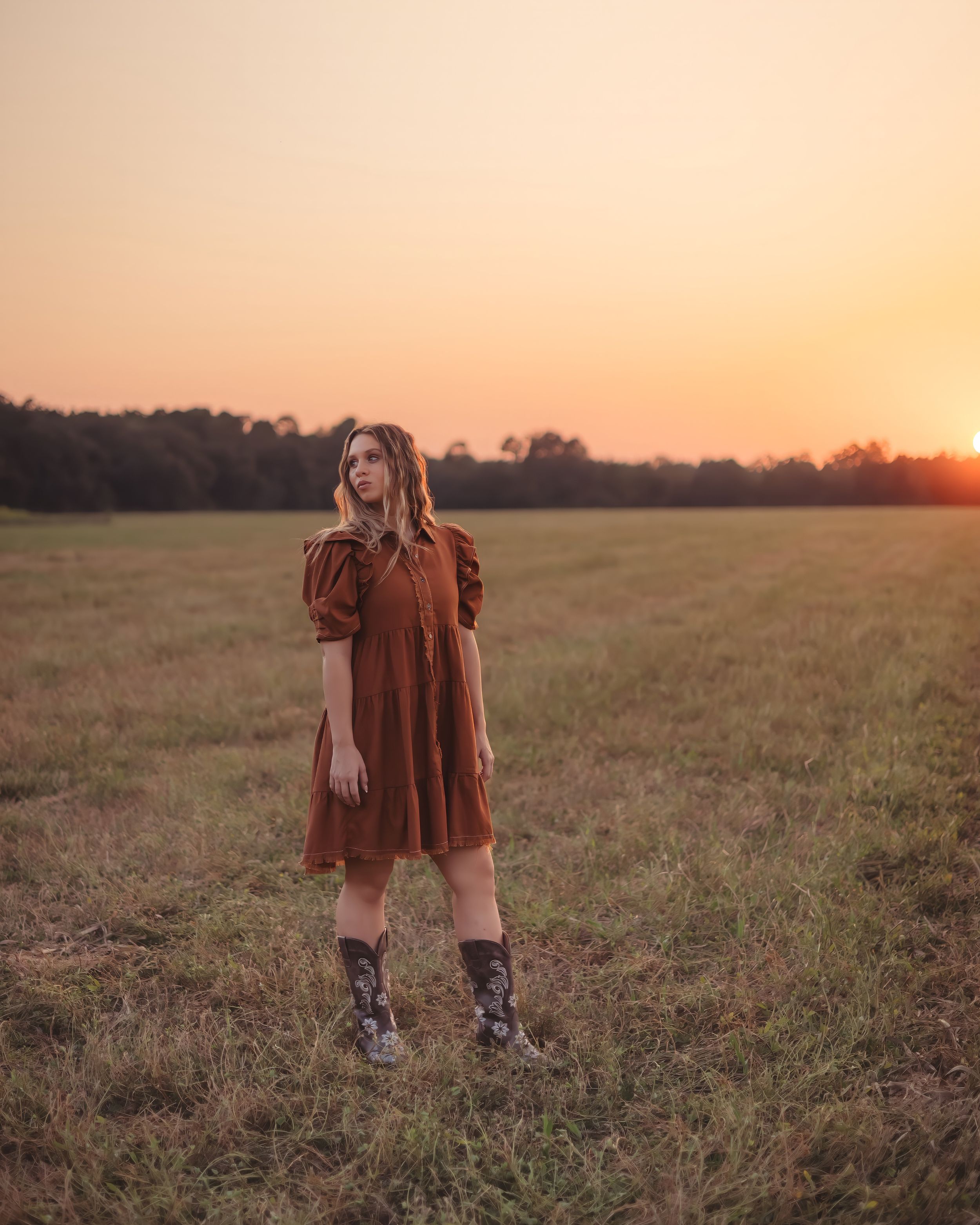 Young woman in brown puff-sleeve dress and cowboy boots gazes upward in open field at sunset.