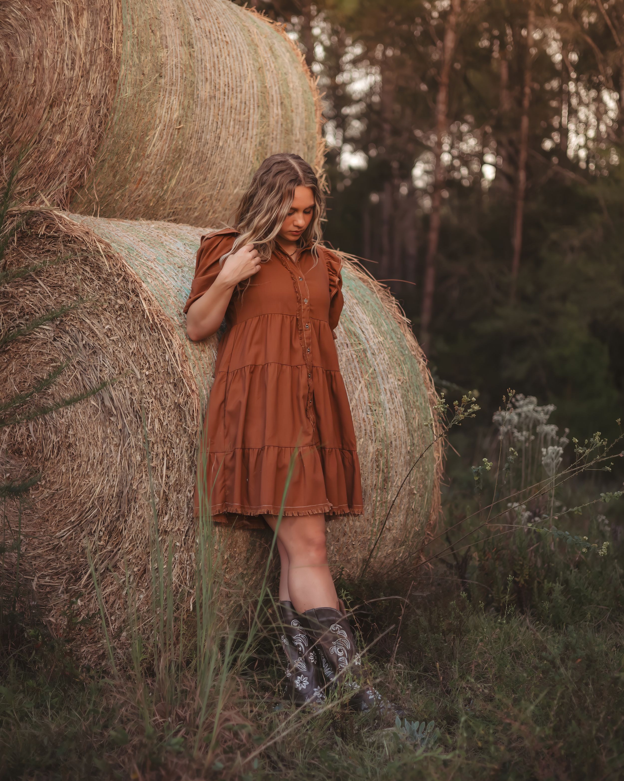 Young woman in rust brown dress leans against large hay bale in wooded farm setting.