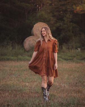 Smiling young woman in rust dress twirls near hay bale in rustic farm field at golden hour.