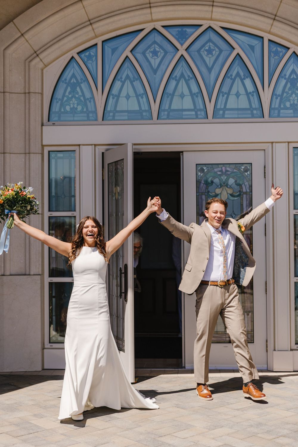 Joyful newlyweds celebrate outside church doors, bride in white gown holding bouquet, groom in tan suit, arms raised.