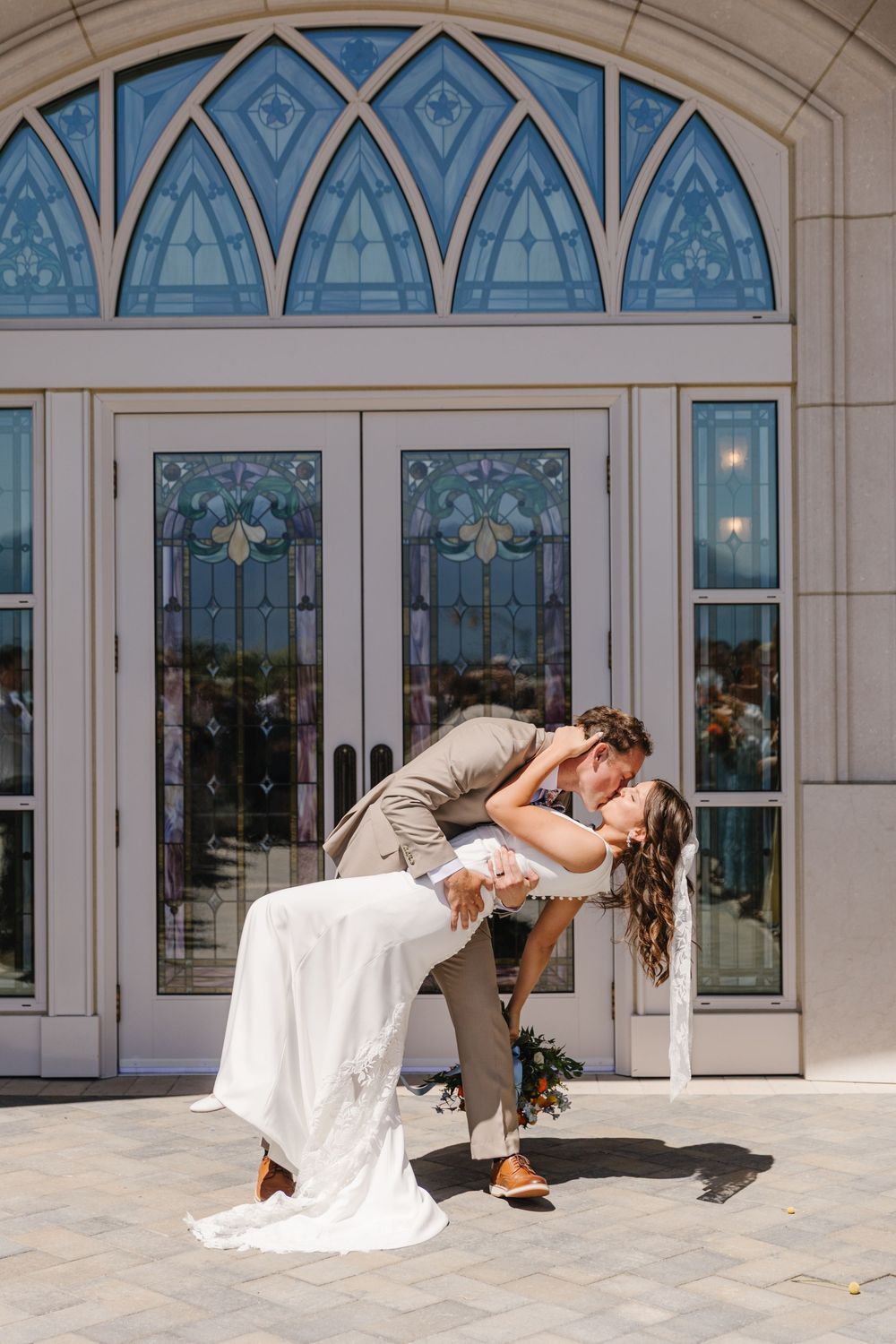 Groom dips bride in romantic kiss outside church with ornate stained glass windows on sunny wedding day.
