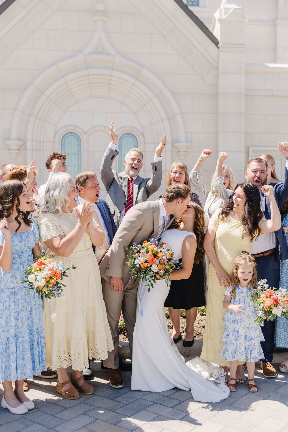 Wedding party celebrates outside white church as groom kisses bride in white dress holding colorful orange bouquet.