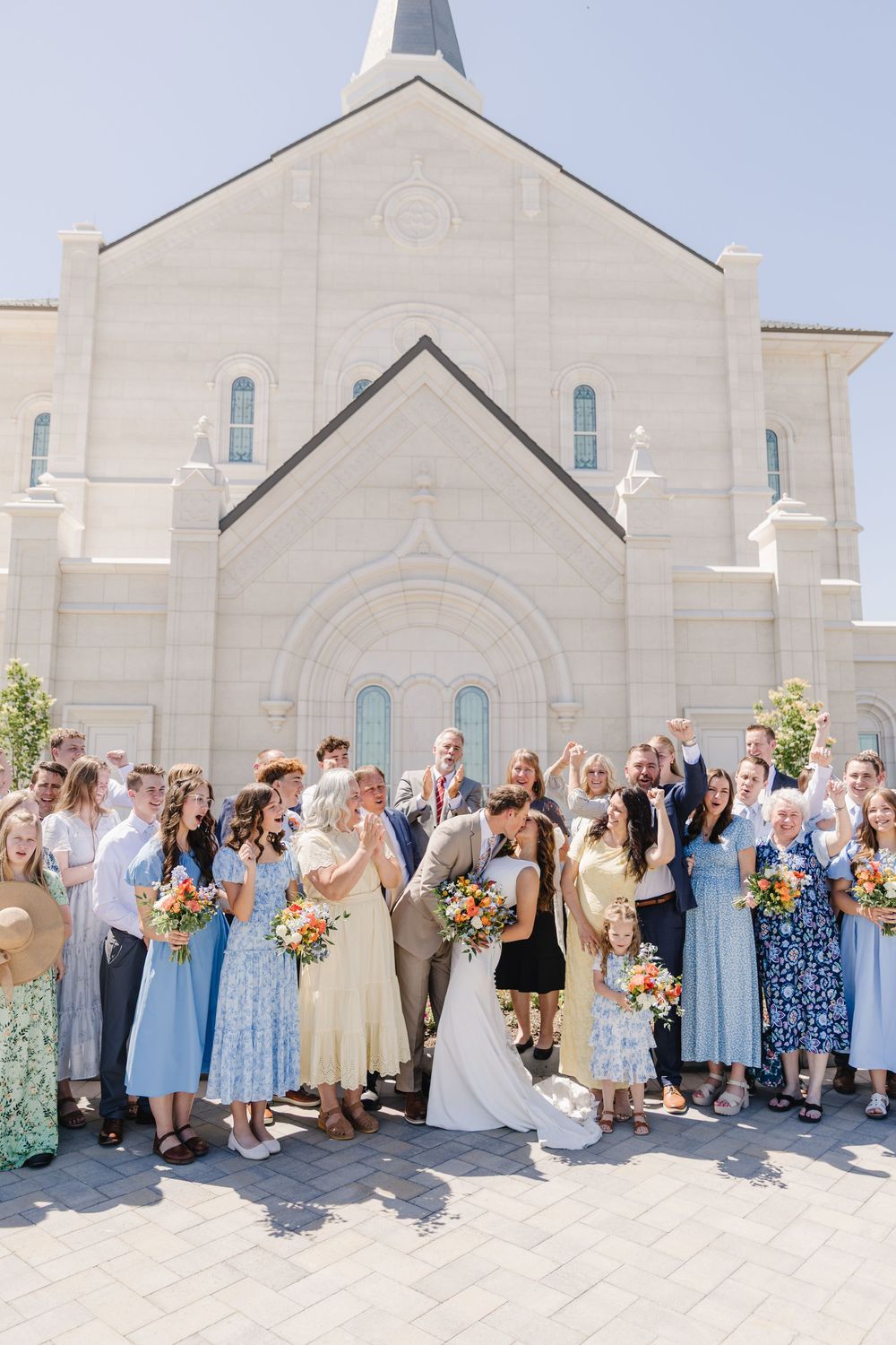 Wedding party celebrates outside a white stone church on a sunny day, bride and groom surrounded by guests in blue dresses.