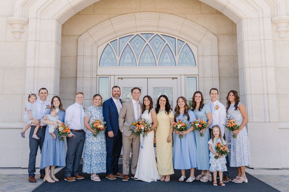 Large wedding party poses in blue attire outside a white stone LDS temple entrance.