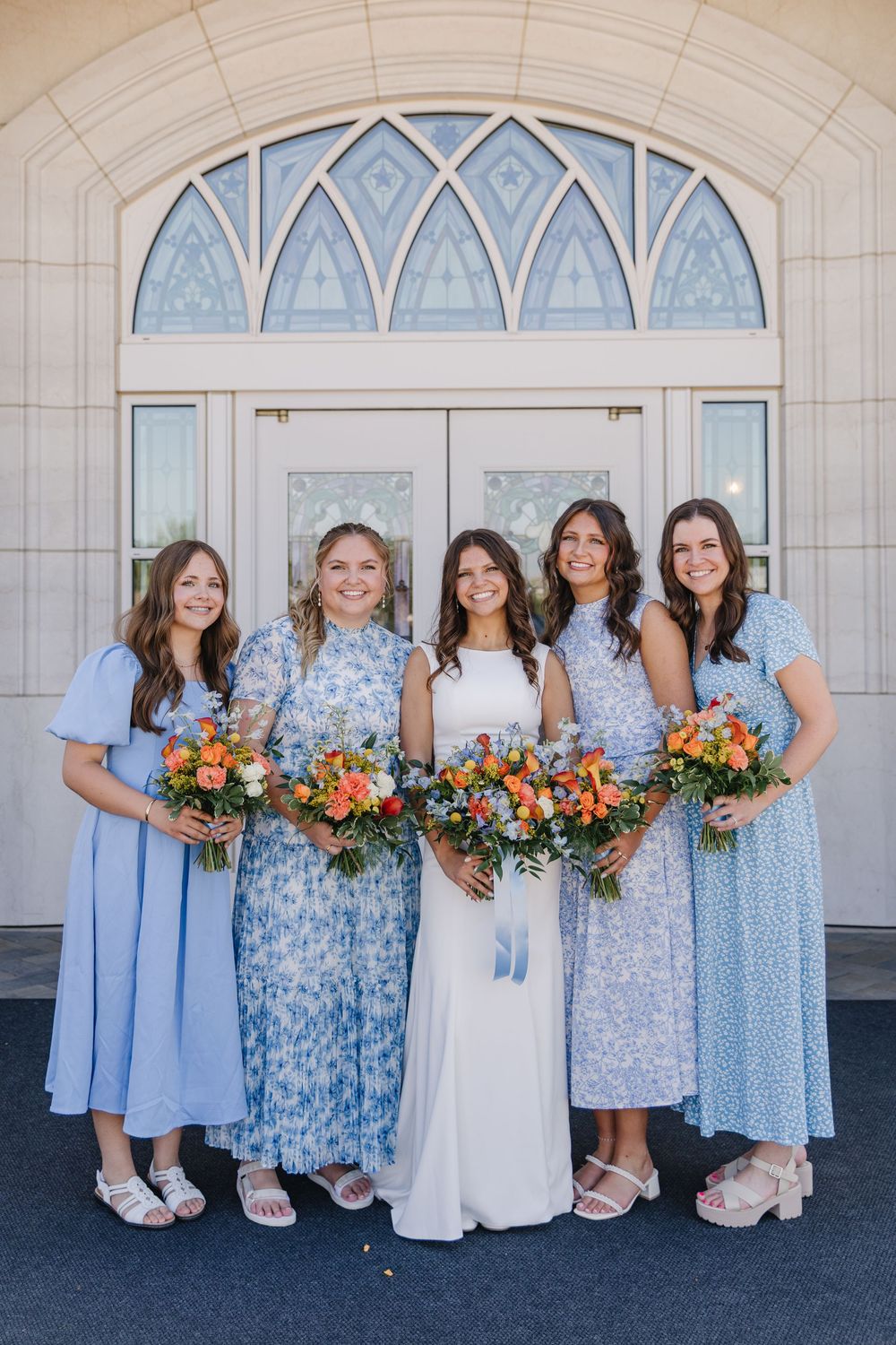 Bride in white gown poses with four bridesmaids in blue dresses holding colorful bouquets outside a church.