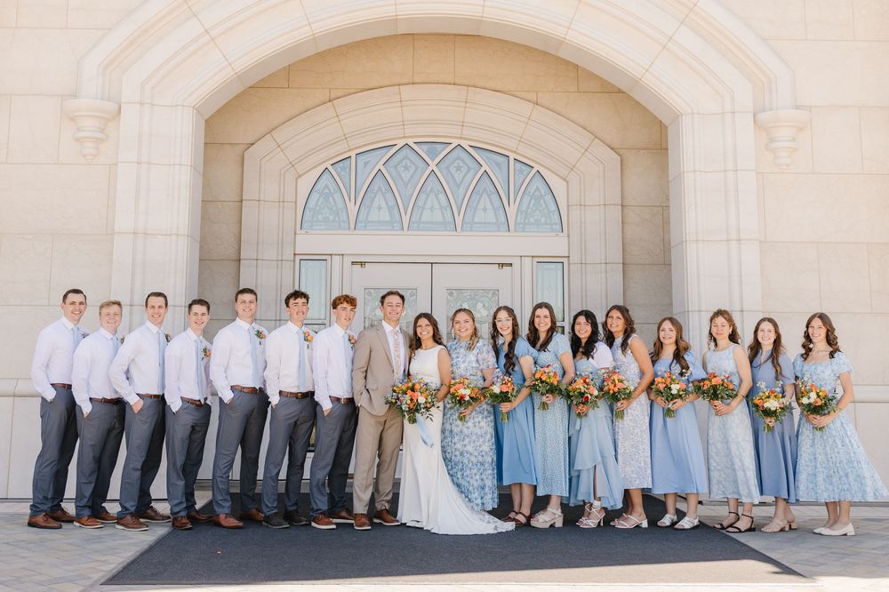 Wedding party poses in blue dresses and gray suits outside a white stone church.