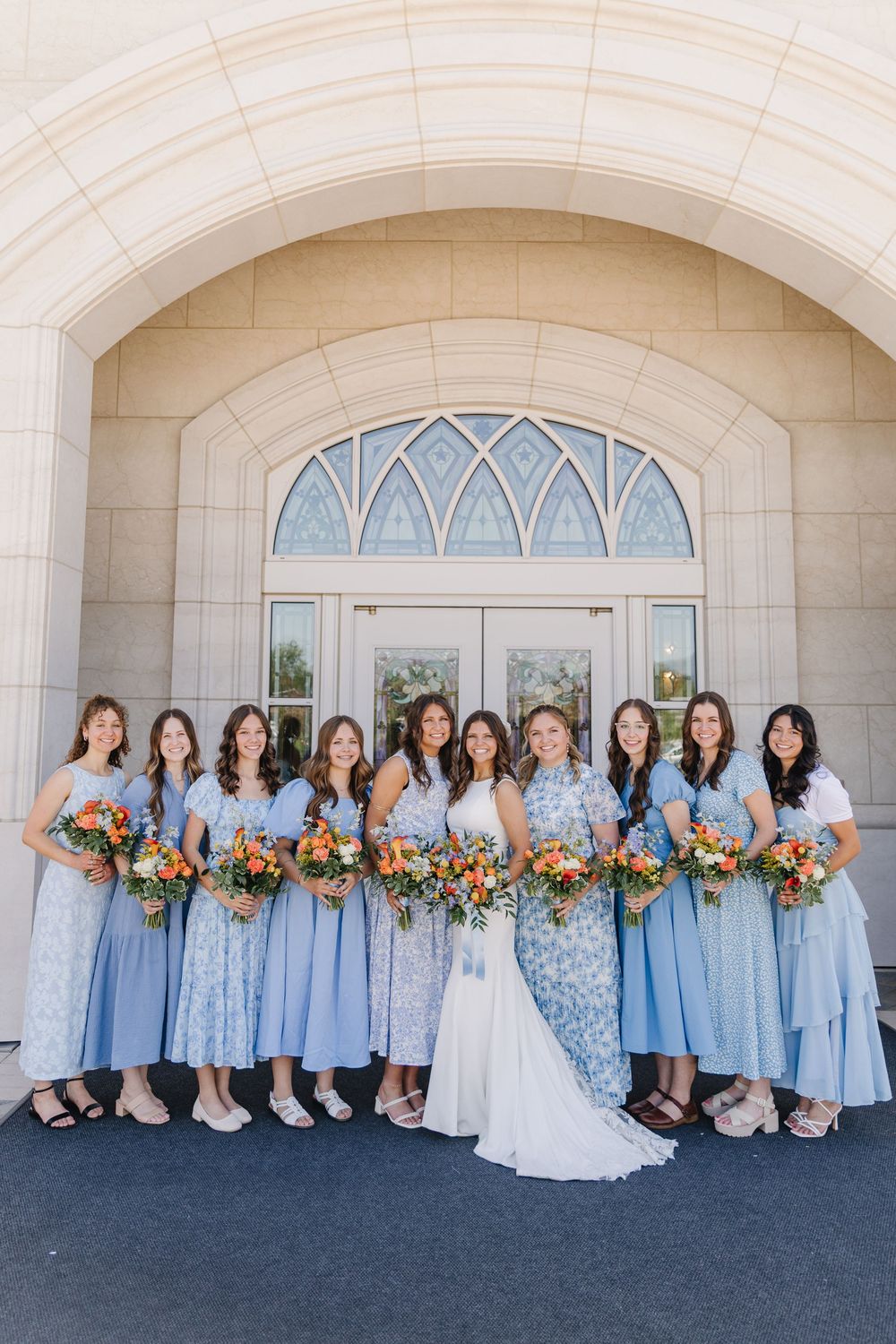 Bride poses with ten bridesmaids in matching light blue dresses holding colorful bouquets outside church.