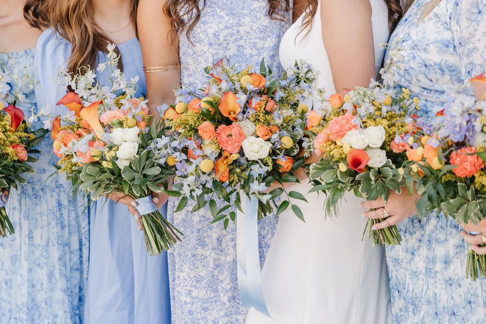 Close-up of bride and bridesmaids holding vibrant orange, peach, and white wedding bouquets with greenery.