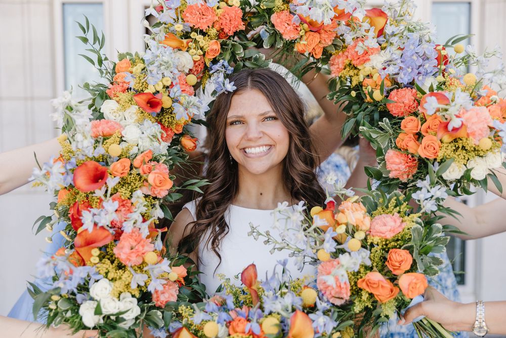 Smiling bride framed by large circular floral arrangement of orange, white, and blue wedding flowers outdoors.