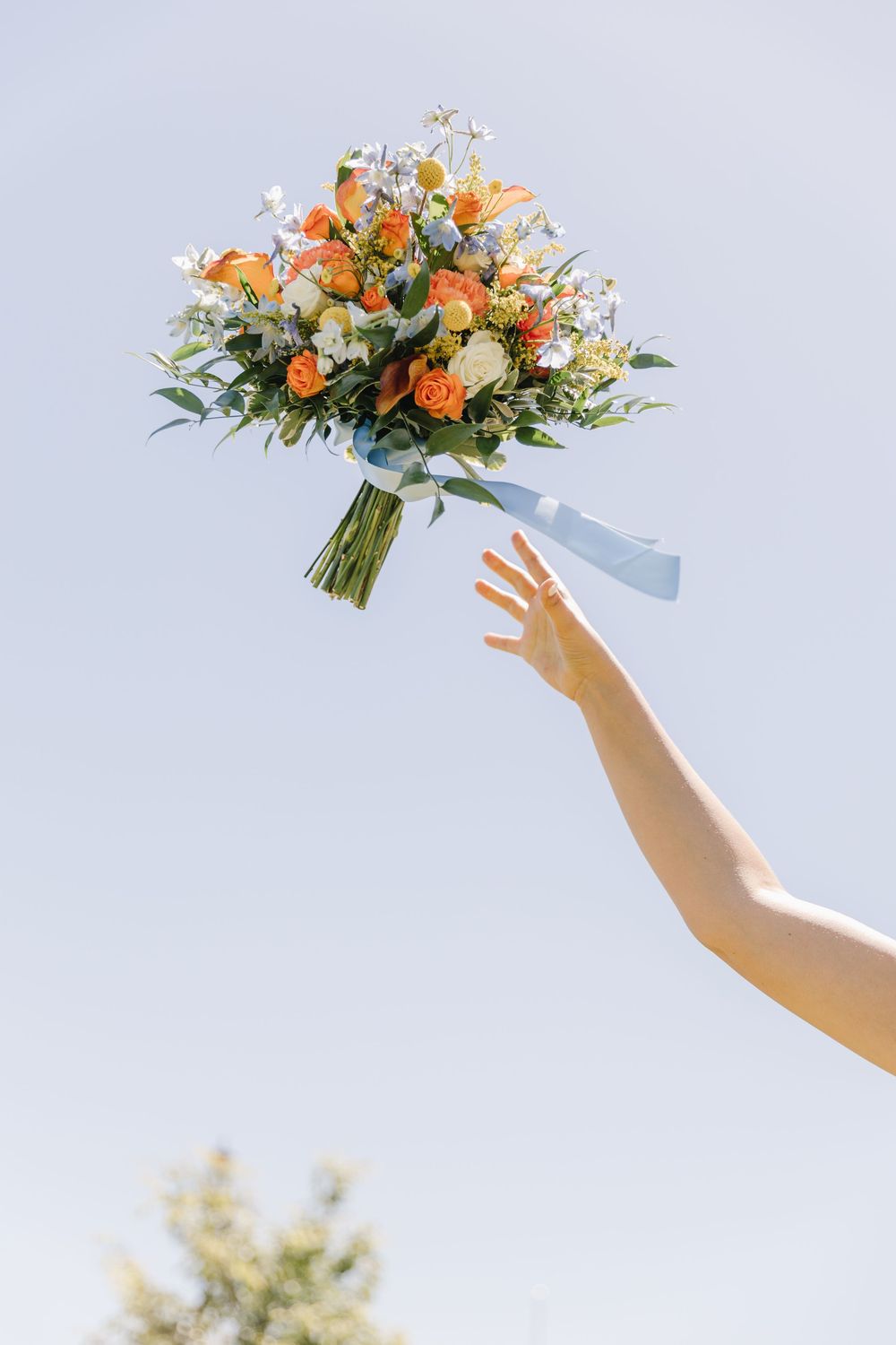 Bride tosses colorful orange and white wedding bouquet with blue ribbon into clear blue sky outdoors.