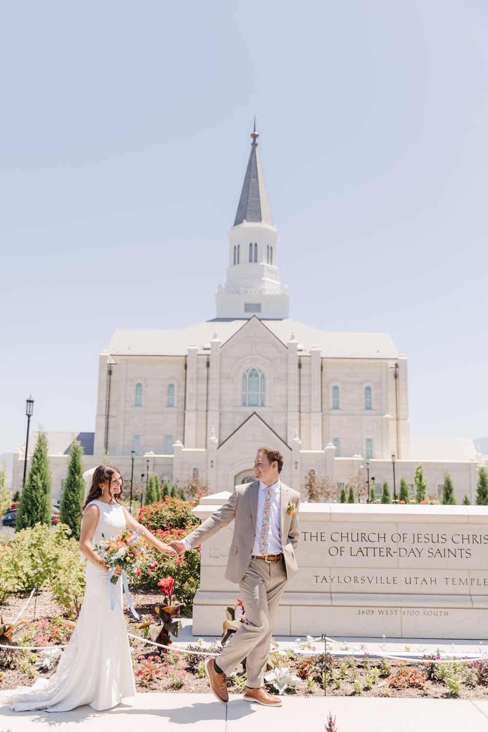 Bride and groom walk hand-in-hand before Taylorsville Utah LDS Temple on sunny day.
