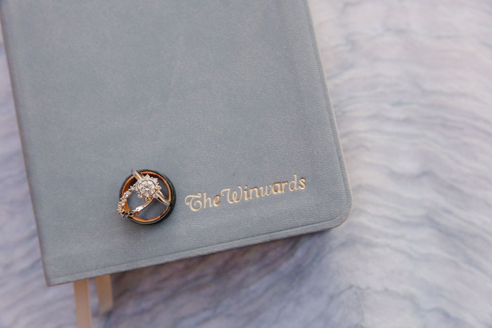 Wedding rings rest on gray velvet journal embossed with The Winwards on marble surface.