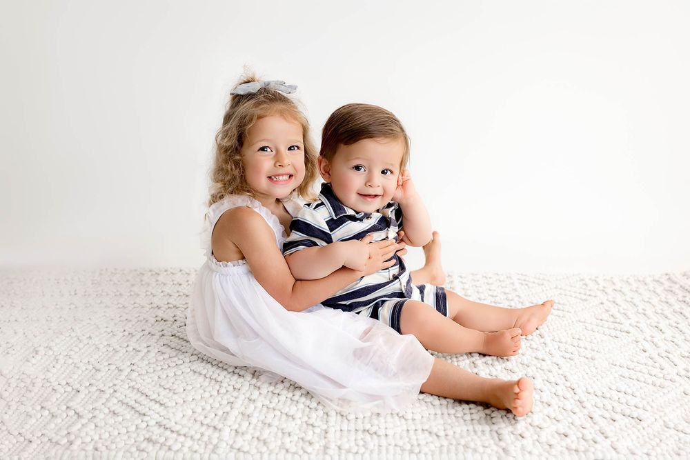 Two young siblings sit together on a white rug, smiling, girl in white dress hugging baby boy in navy striped outfit.