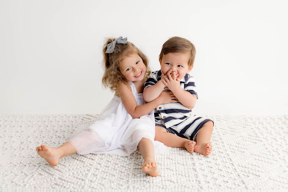 Two young siblings sitting together on a white textured blanket, smiling and playing in matching white and navy outfits.