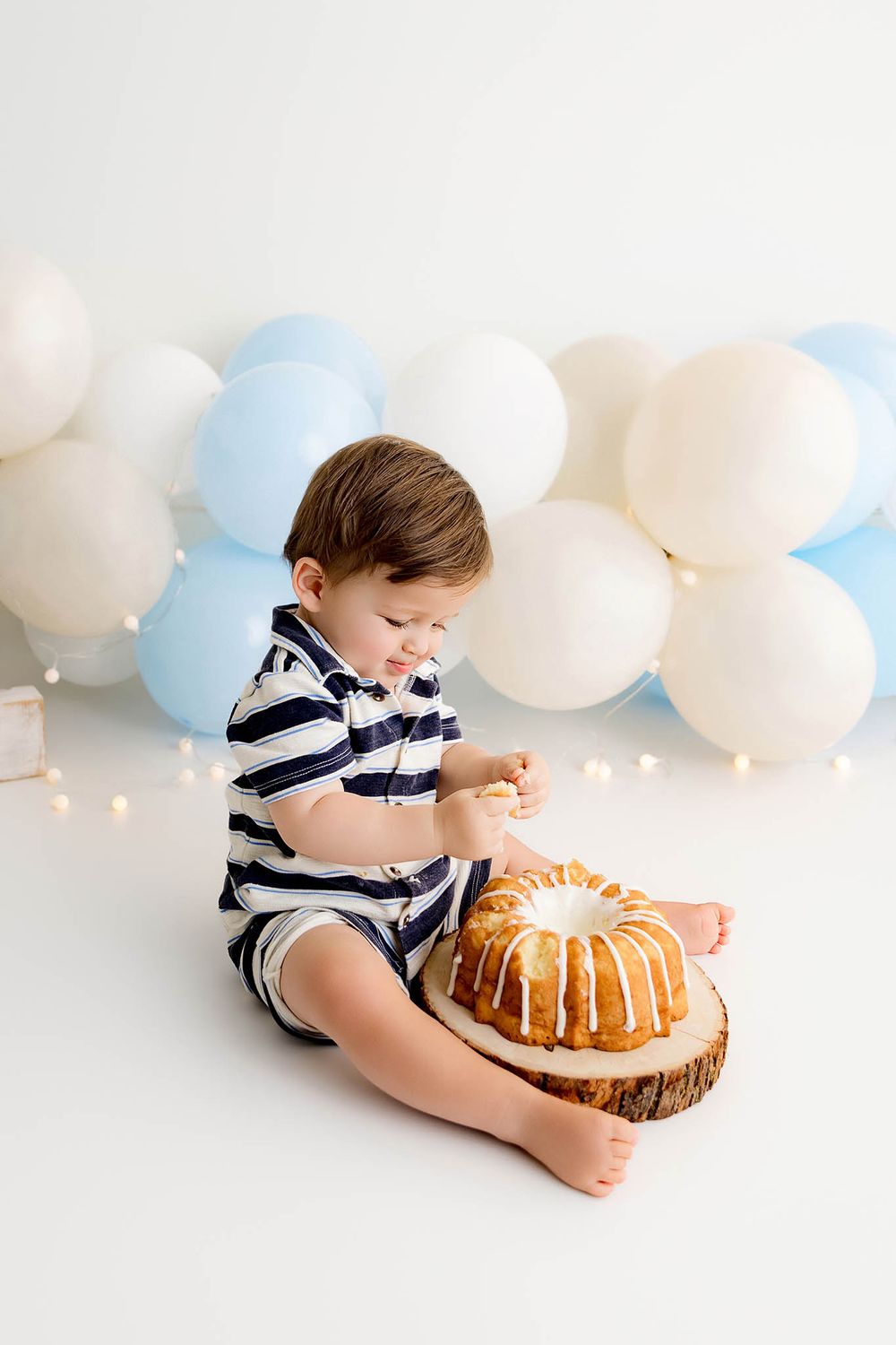 Toddler boy in striped romper sitting on floor exploring birthday bundt cake with white balloons backdrop.