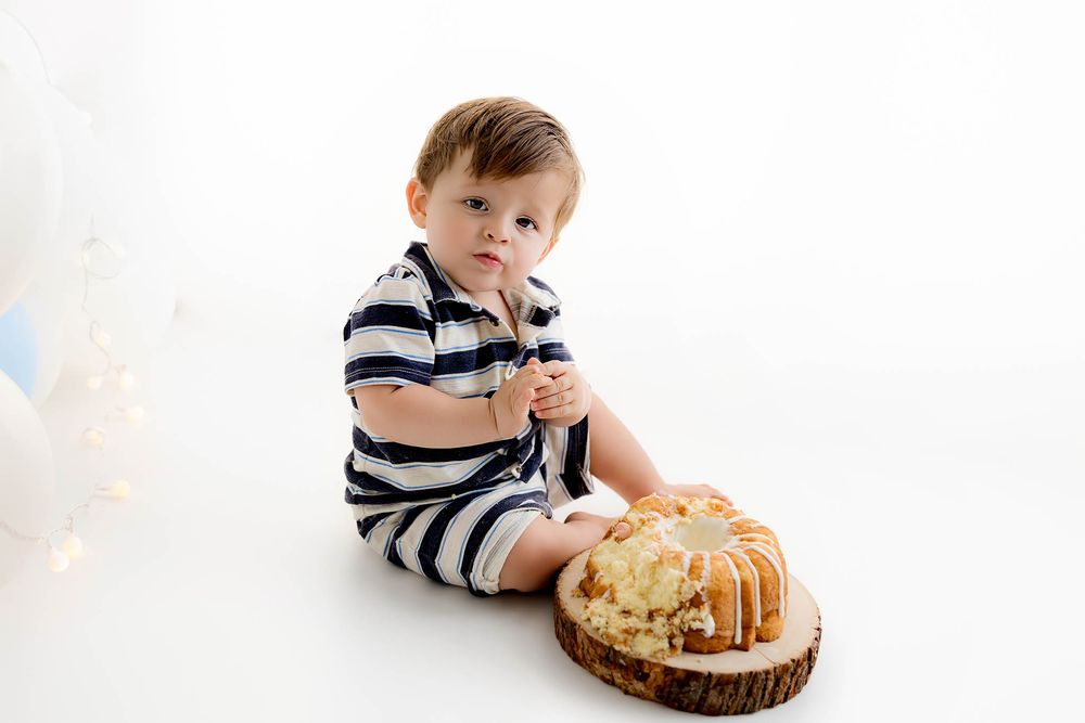 Baby boy in striped romper sits on white backdrop beside smashed first birthday cake on wooden slice.
