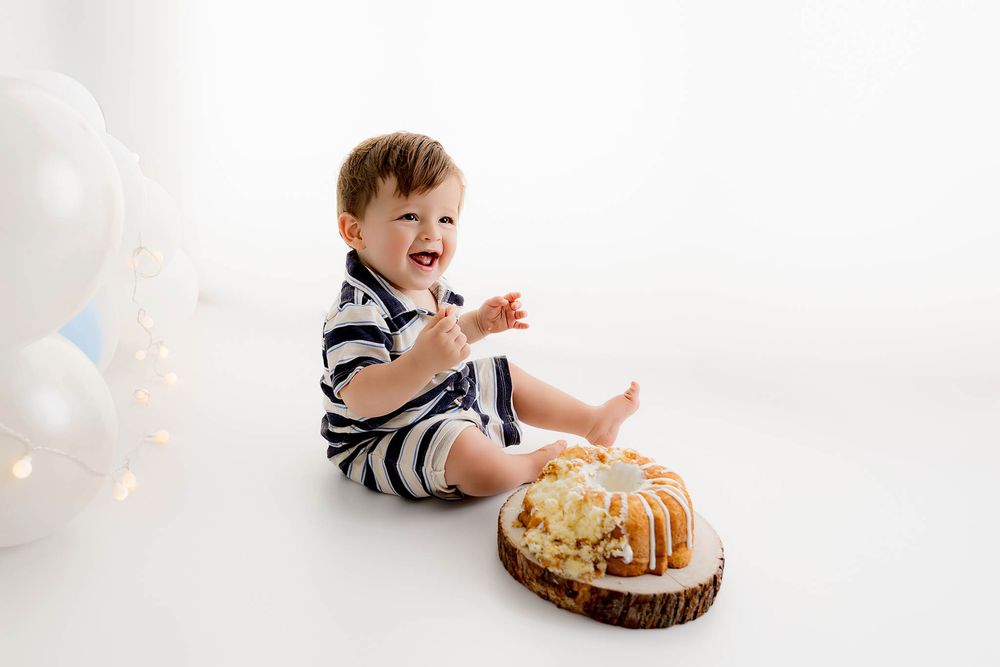 Happy baby boy in striped romper laughing during first birthday cake smash photo session with white balloons.
