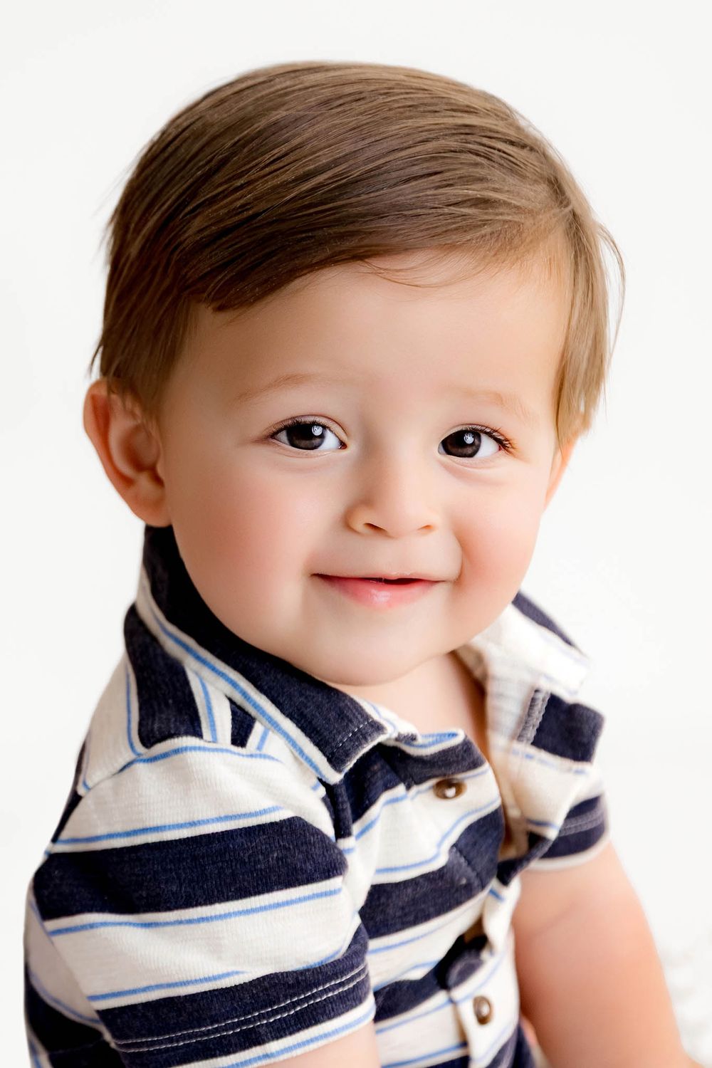Adorable toddler boy with brown eyes smiling, wearing a navy striped polo shirt, portrait photo on white background.