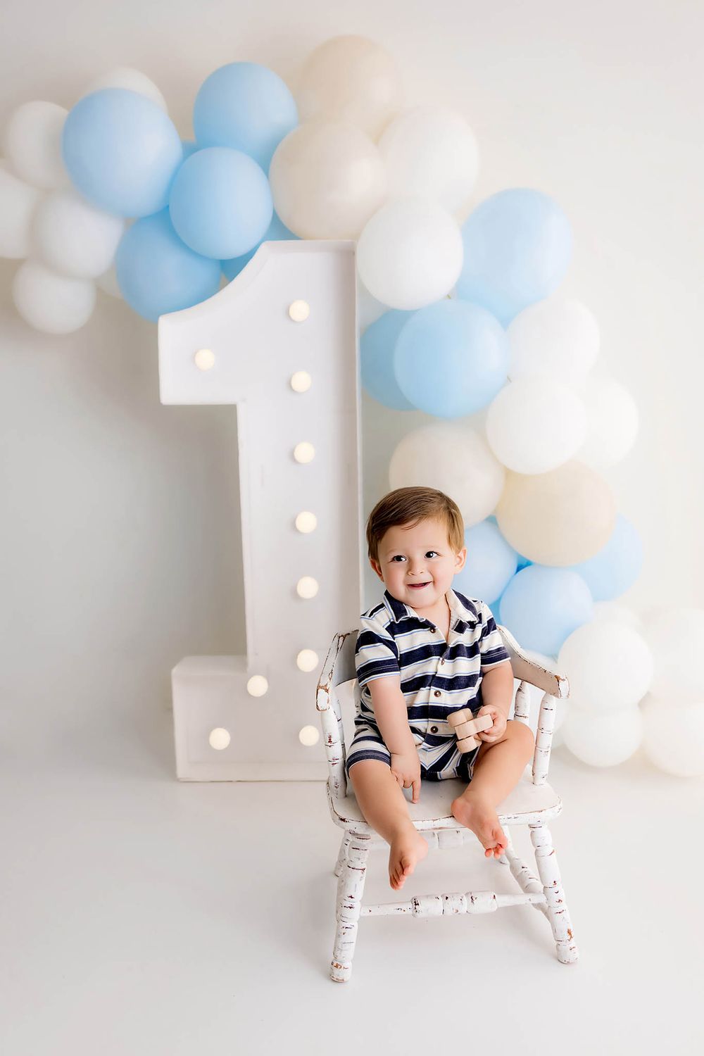 Smiling baby boy sitting in white chair beside illuminated number 1 with blue and white balloon garland.