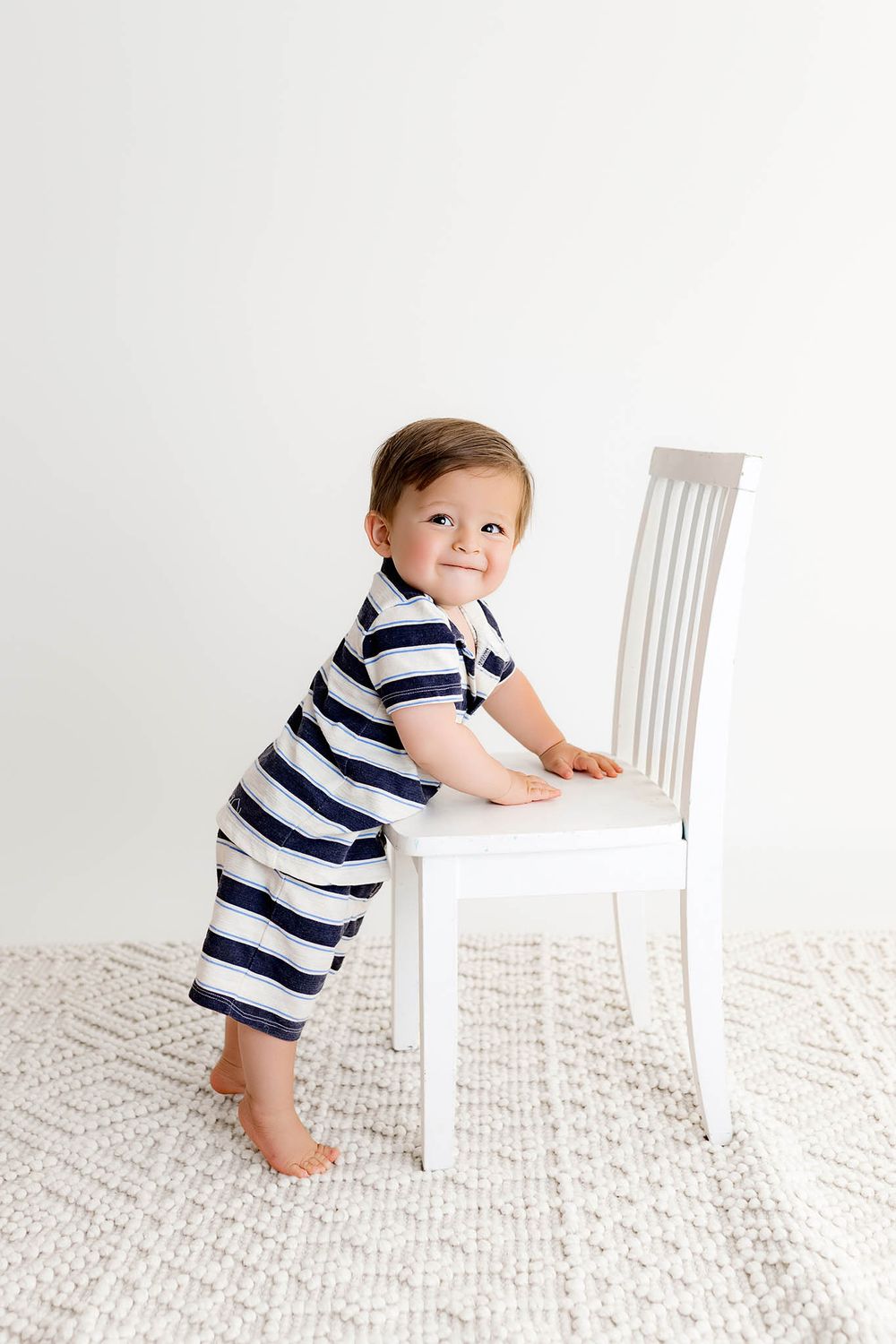 Smiling baby boy in navy striped romper standing barefoot beside a white chair on a textured rug.