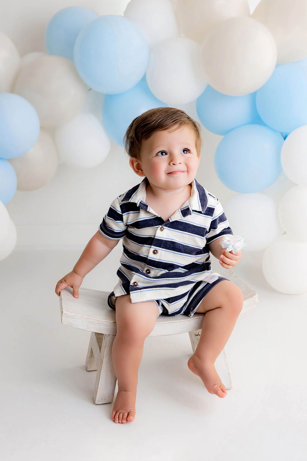 Smiling toddler boy in striped romper sitting on white bench with blue and white balloon backdrop.