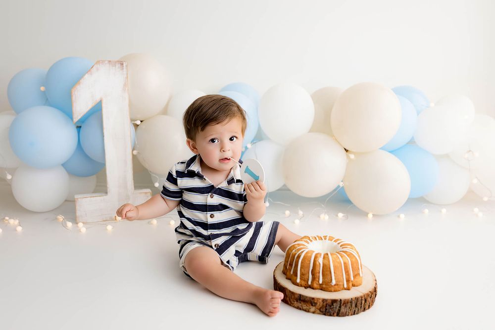 Baby boy in striped romper sits by bundt cake with blue and white balloons and number one for first birthday smash.