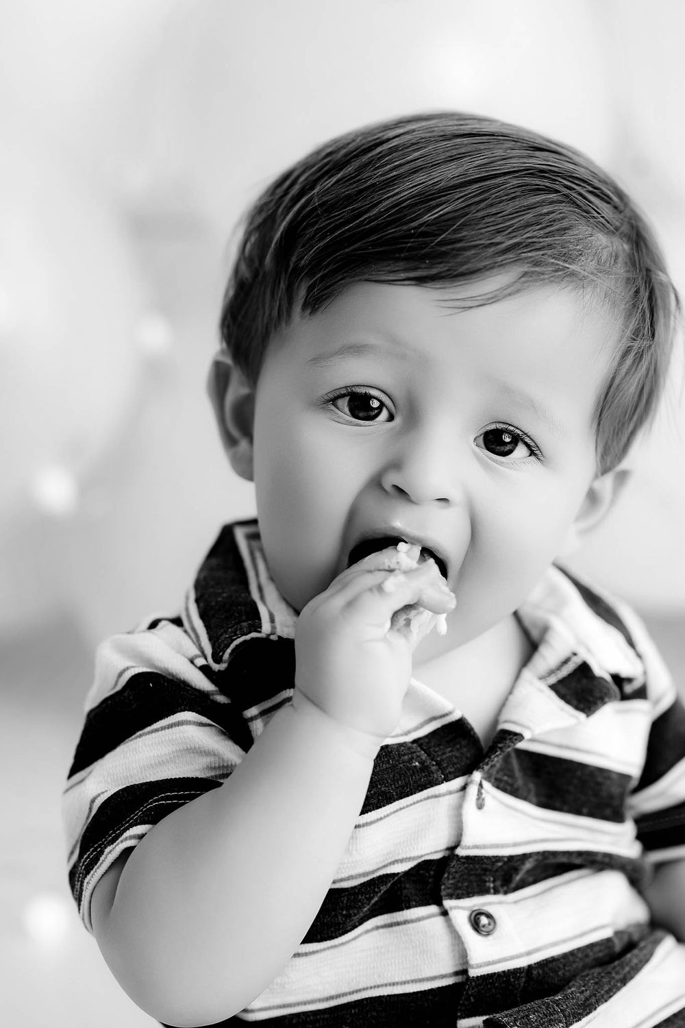 Black and white portrait of a cute toddler boy in a striped shirt eating cake for first birthday photoshoot.