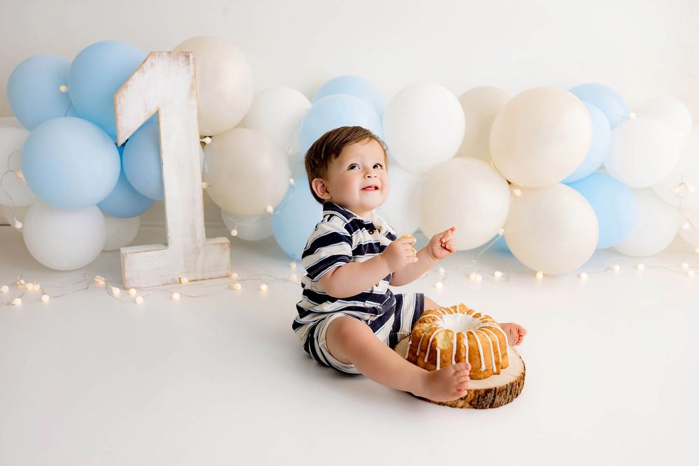 Adorable baby boy celebrating his first birthday cake smash surrounded by blue and white balloons.