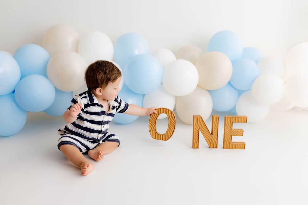 Baby boy in striped romper sits by wooden ONE letters with blue and white balloon garland for first birthday photo.
