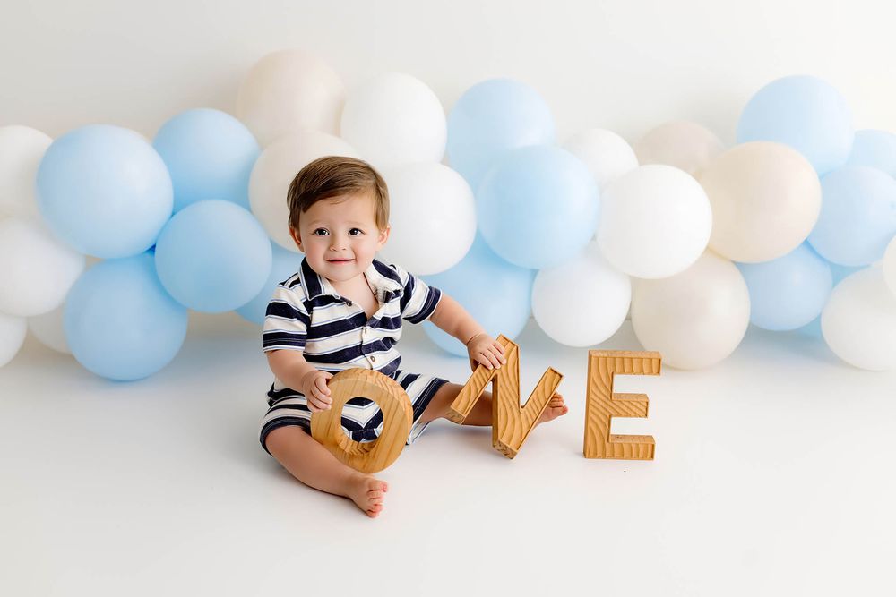 Smiling baby boy in striped romper sitting with wooden ONE letters during first birthday photo session.