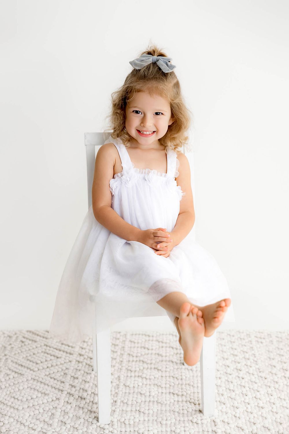 Adorable smiling little girl in white dress with gray bow sitting barefoot on white chair against white wall.