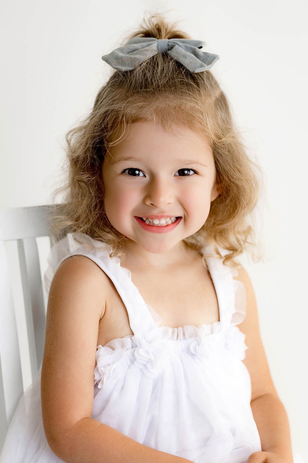 Smiling young girl with curly blonde hair wearing a white dress and gray bow, posed for a portrait photo.