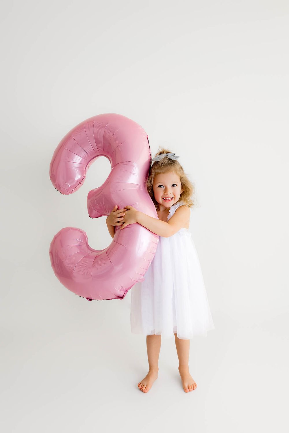 A smiling little girl in a white tutu dress hugging a large pink number 3rd birthday balloon.