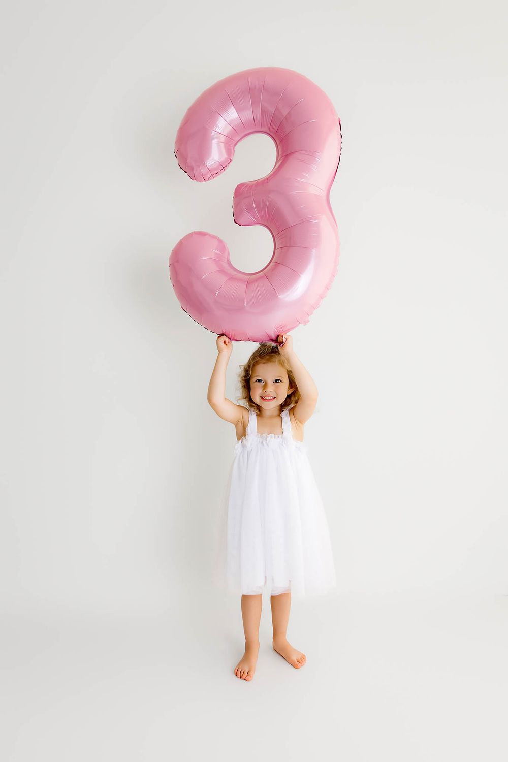 A happy little girl in a white dress holding a large pink number 3rd birthday balloon above her head.