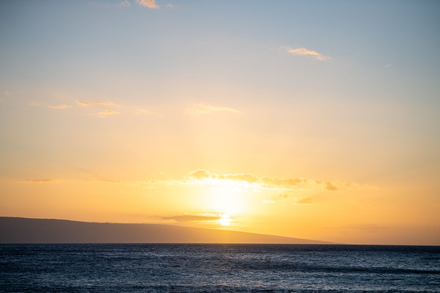 Golden sunset over calm ocean waters with silhouetted island on horizon and soft clouds glowing orange.