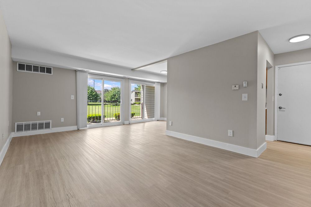 Bright empty living room with light wood floors, gray walls, sliding glass door to balcony, and white trim.