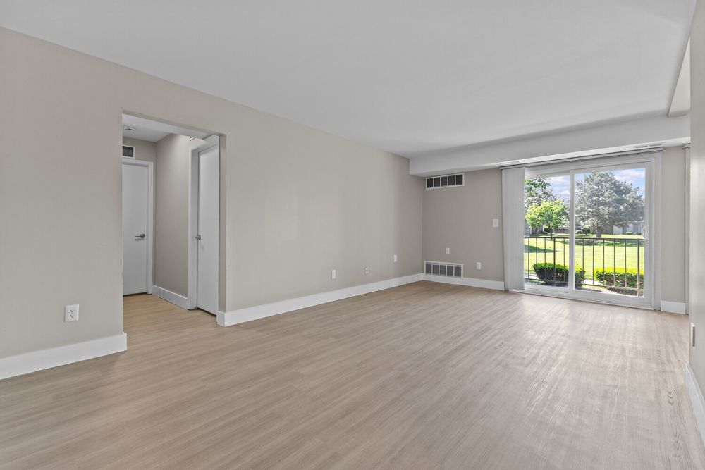 Bright empty living room with light hardwood floors, large sliding glass door, and neutral walls leading to a hallway.