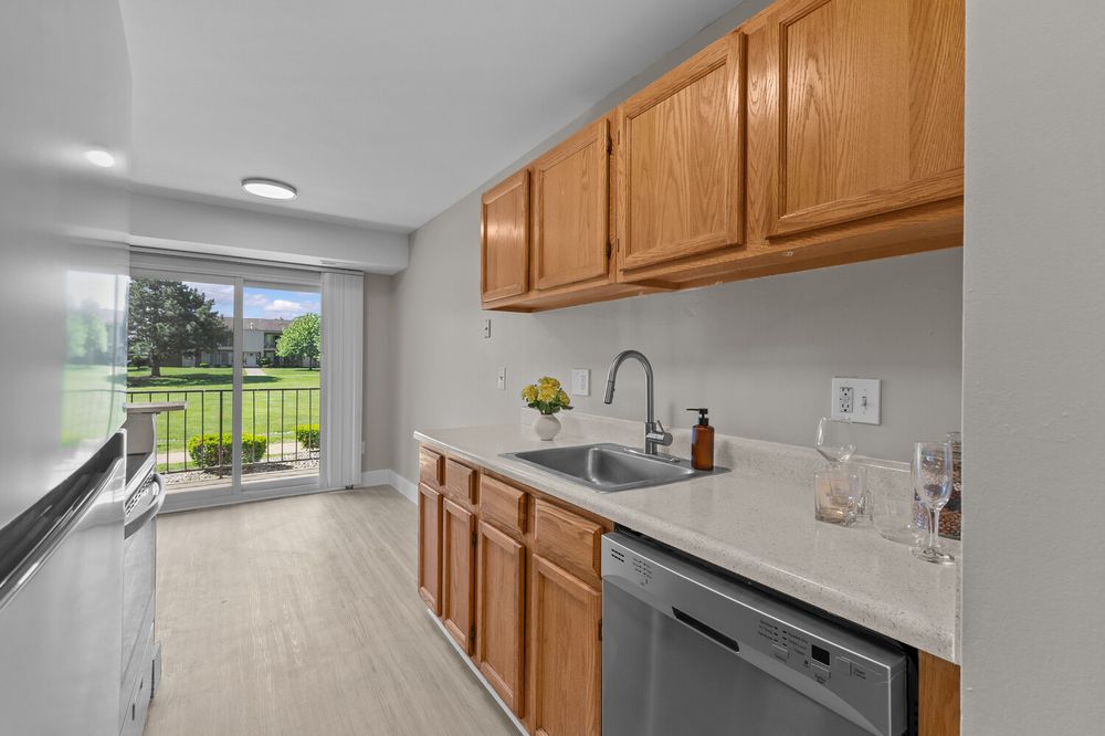 Modern kitchen with oak cabinets, stainless steel dishwasher, and sliding glass door overlooking green backyard.