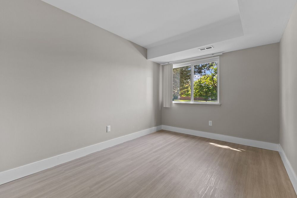 Bright empty room with light wood floors, neutral gray walls, and a window with natural light.